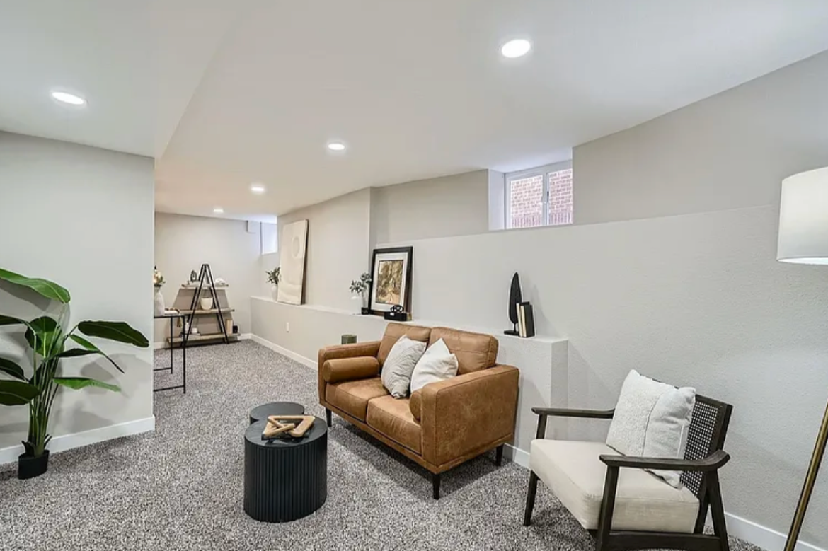Modern living room with a tan sofa, white armchair, black coffee table, potted plant, artwork, and recessed lighting.