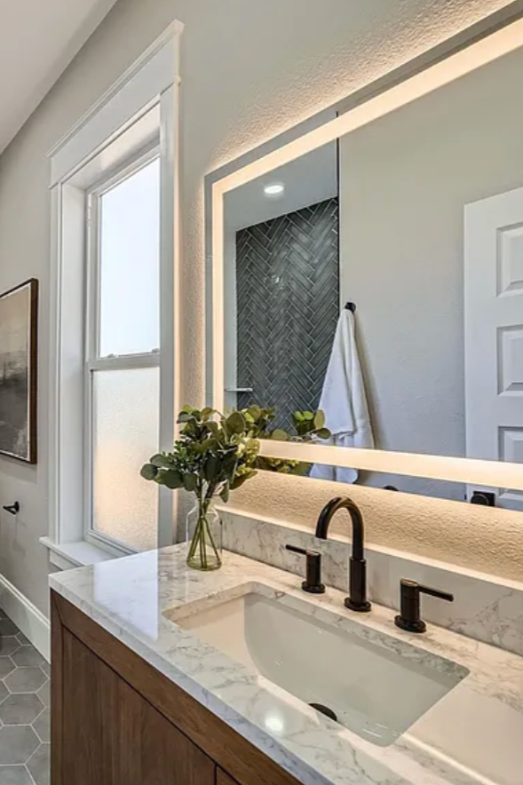 Bathroom with a vanity featuring a marble countertop, a vessel sink, and wall-mounted black faucet fixtures. There is a mirror with built-in lighting above the sink, a window with white trim, and a vase with greenery on the countertop. A towel is hanging inside a shower area with dark herringbone tile, visible behind the mirror.