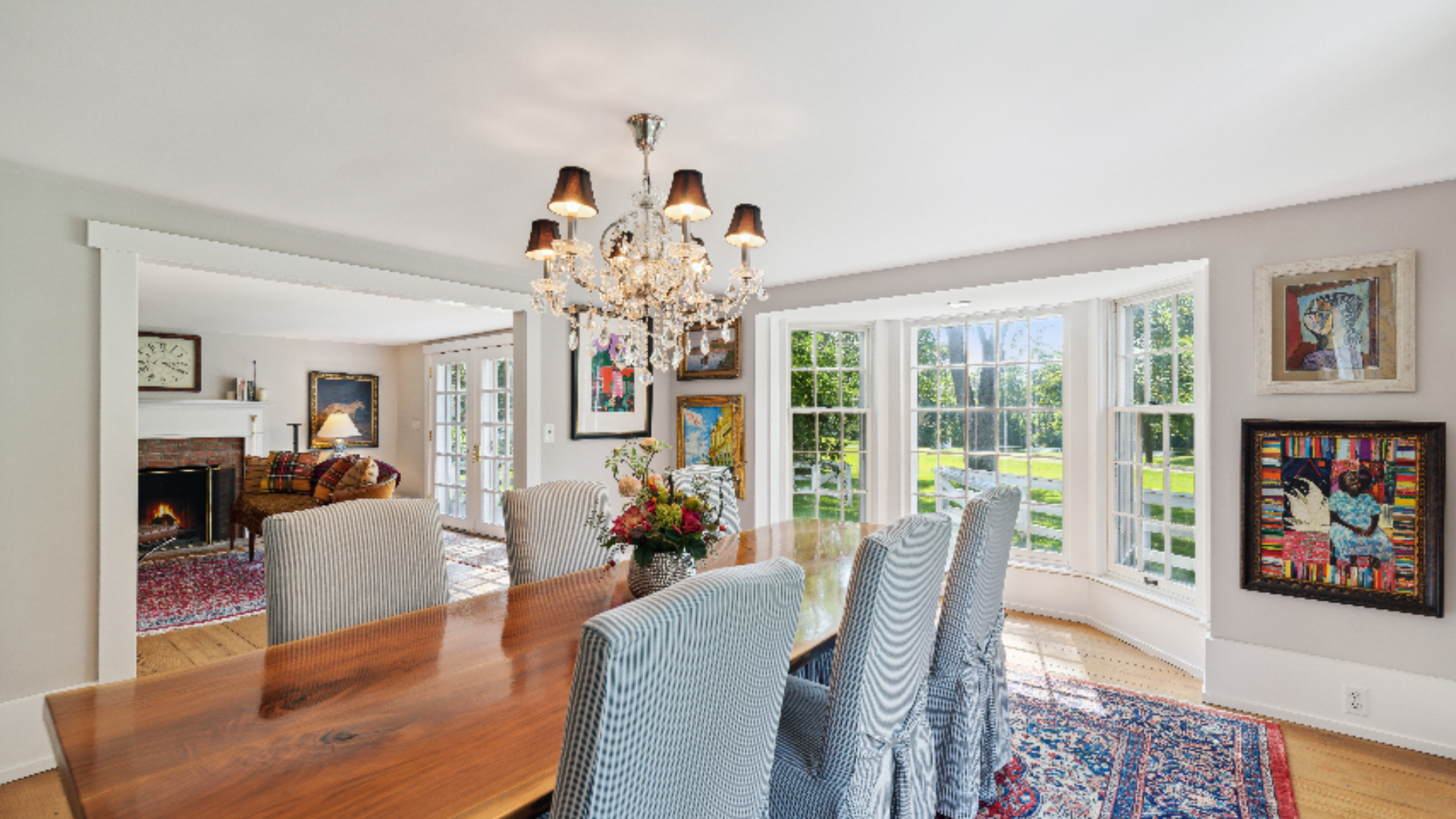 Dining Room Overlook to Living Room - A crystal chandelier sets an elegant tone above the long wood dining table, flanked by upholstered chairs and framed by a bay window with garden views. Bolt contemporary art adds a modern accent against the room'