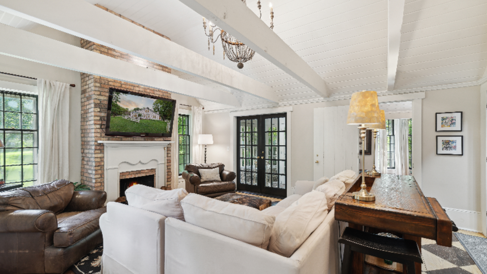 Soaring beamed ceilings anchor this airy living space, centered around a whitewashed brick fireplace and layered with plus white slipcovered sofas alongside a rich leather armchair for contrast. Black-framed French doors and windows flood the room wi