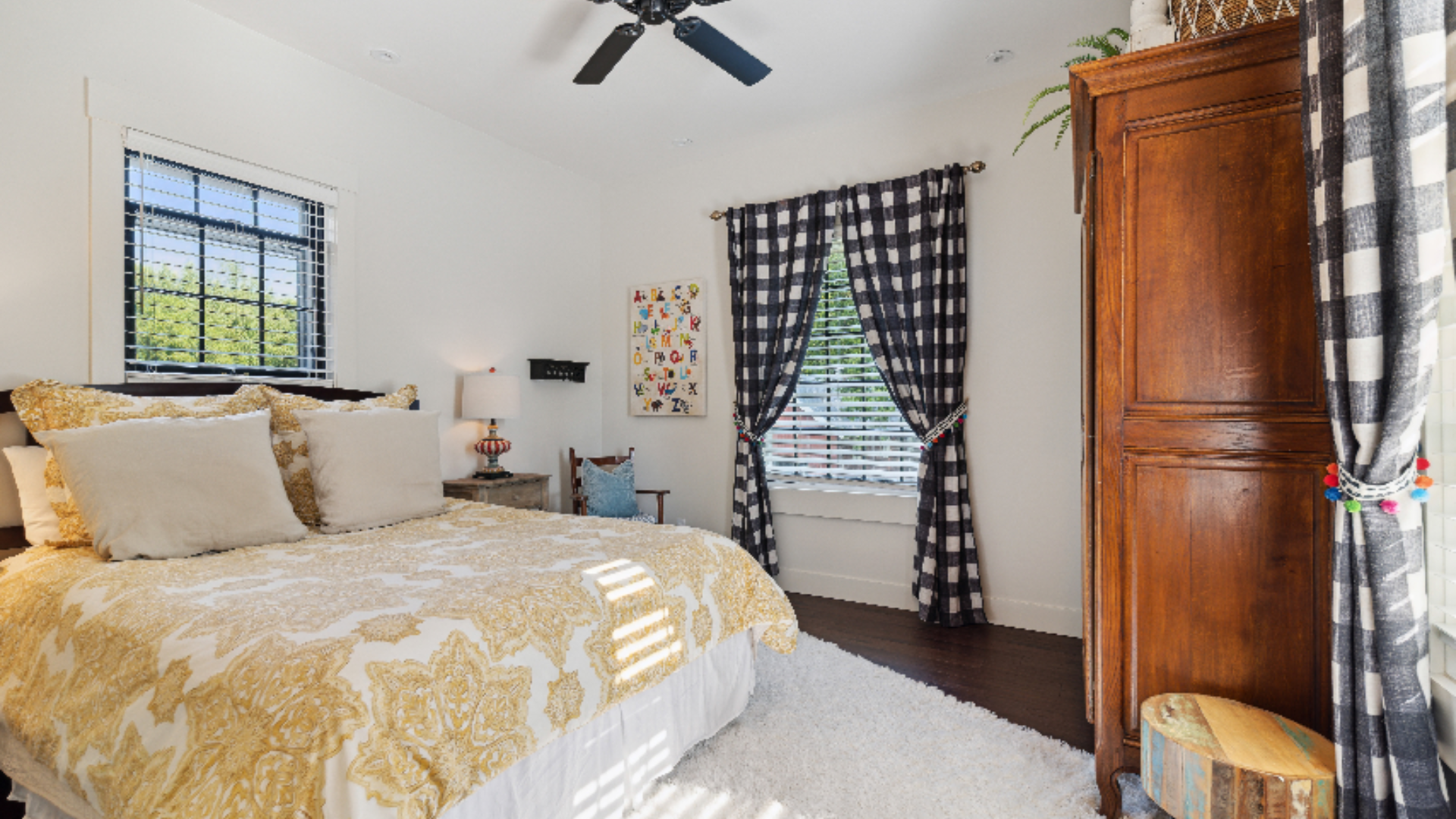 Golden Guest Bedroom - A rich gold damask coverlet brings warmth to this inviting bedroom, paired with crisp white linens and playful navy buffalo-check drapery. An antique wood armoire adds old-world presence, while a plush area rug and soft natural