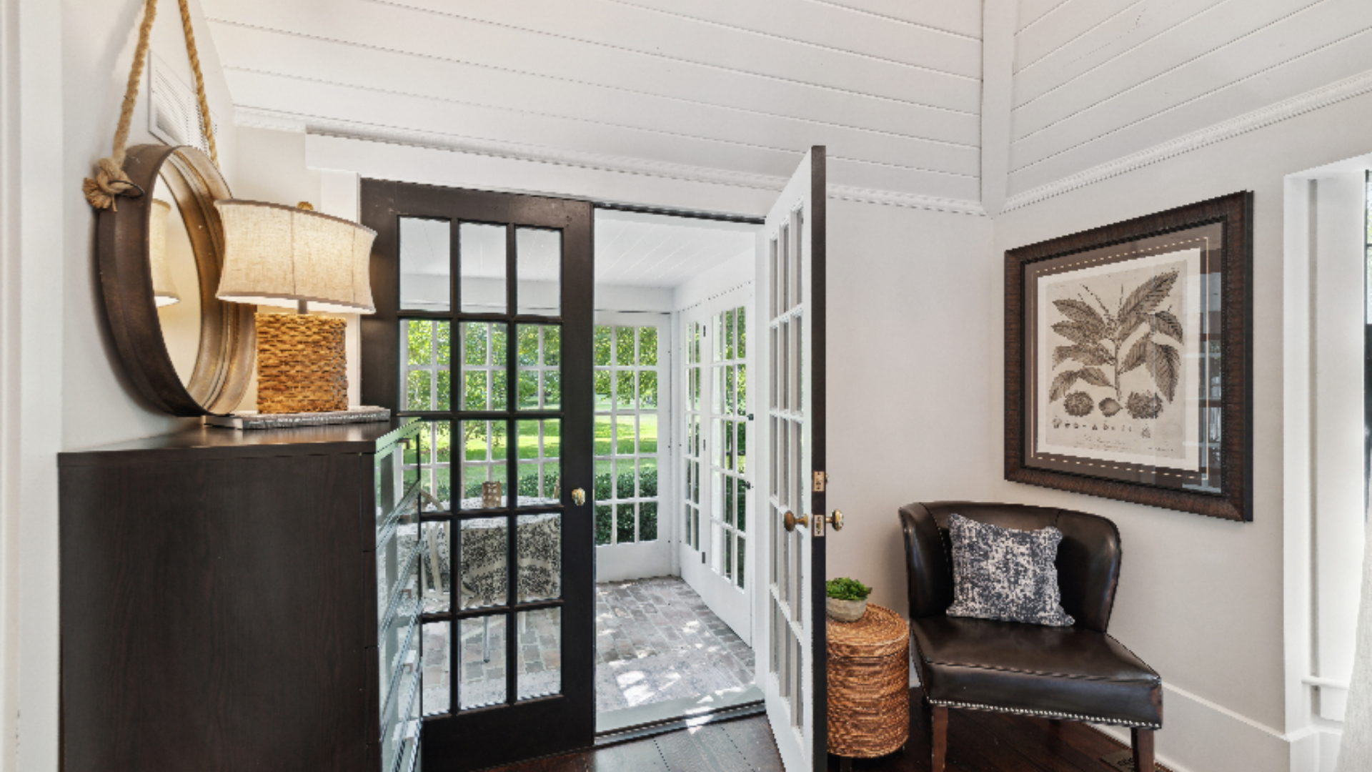 Entry Vignette - A dark chest topped with a hanging pendant lamp and a round mirror creates a striking first impression, opposite a sleek black leather chair and framed botanical print. Black-framed French doors open to the outdoors, blending indoor 