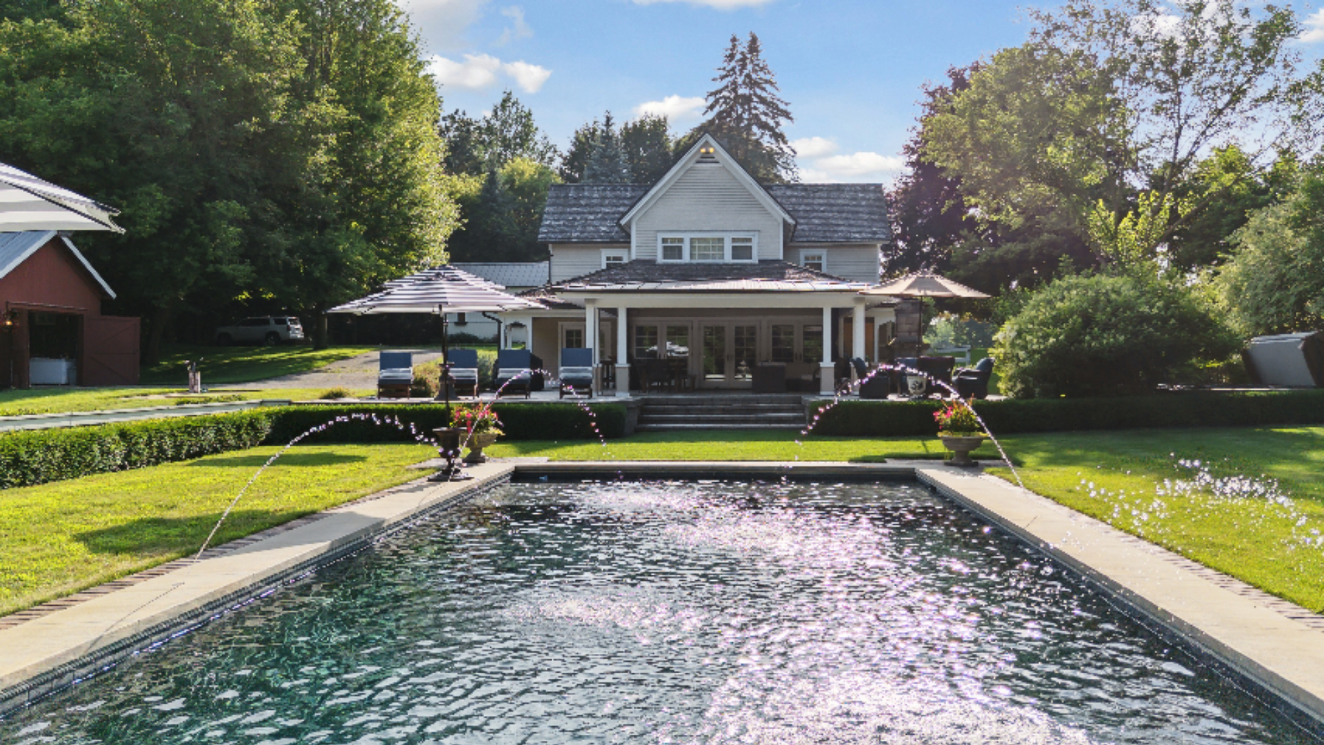 Farmhouse Pool & Grounds - A classic rectangular pool with twin fountain jets sits at the heart of this lush backyard, framed by manicured lawn and mature shade trees. The white farmhouse-style home with its covered travertine veranda offers a pictur