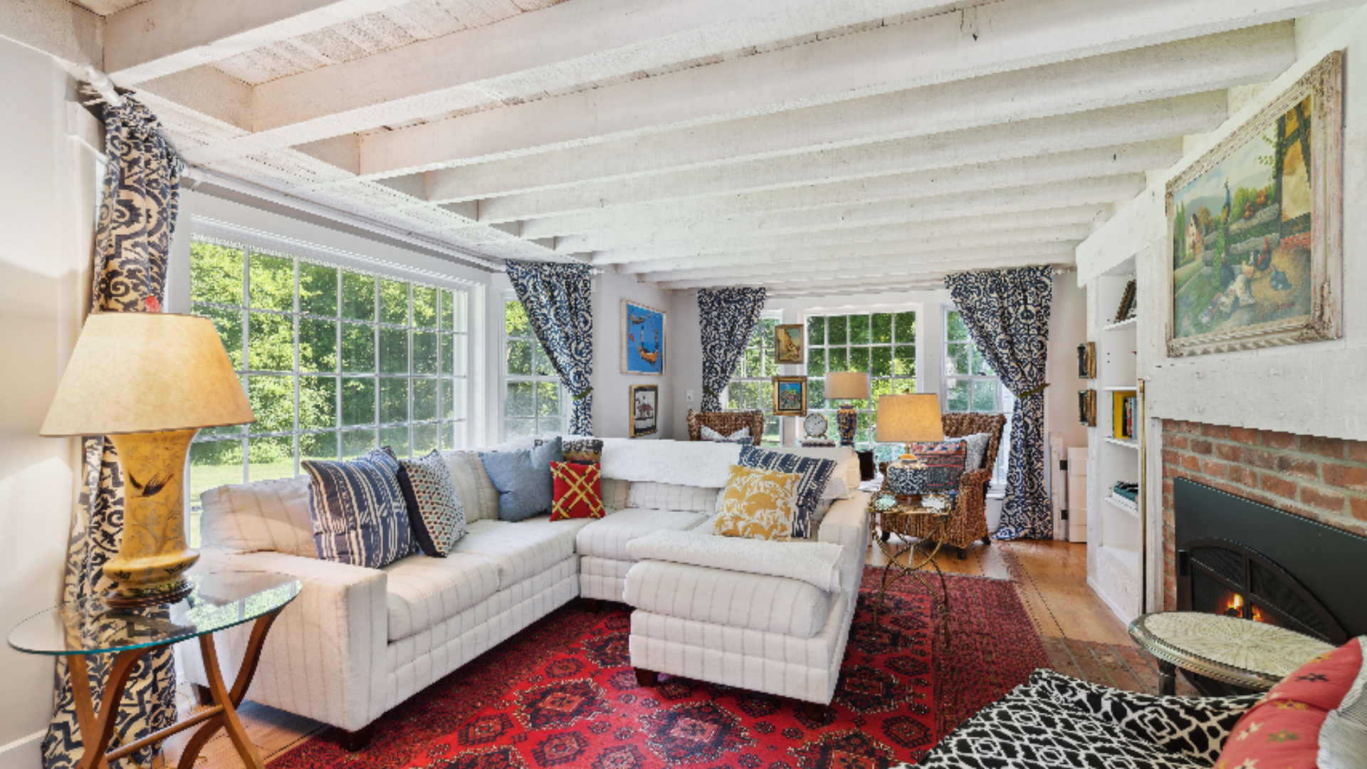 Sun-Filled Sitting Room - A crisp white sectional and matching ottoman offer relaxed seating beneath white-painted beams, grounded by a vidid red patterned rug. Wallls of windows and patterned drapery bring in abundant light, while a brick fireplace 