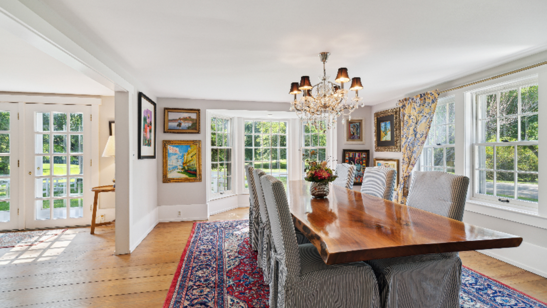 Elegant Dining Room - A live edge wood table anchors this sun-drenched dining space, surrounded by upholstered chairs and grounded by a richly patterned area rug. A crystal chandelier adds a touch of formality, while walls of windows and French doors