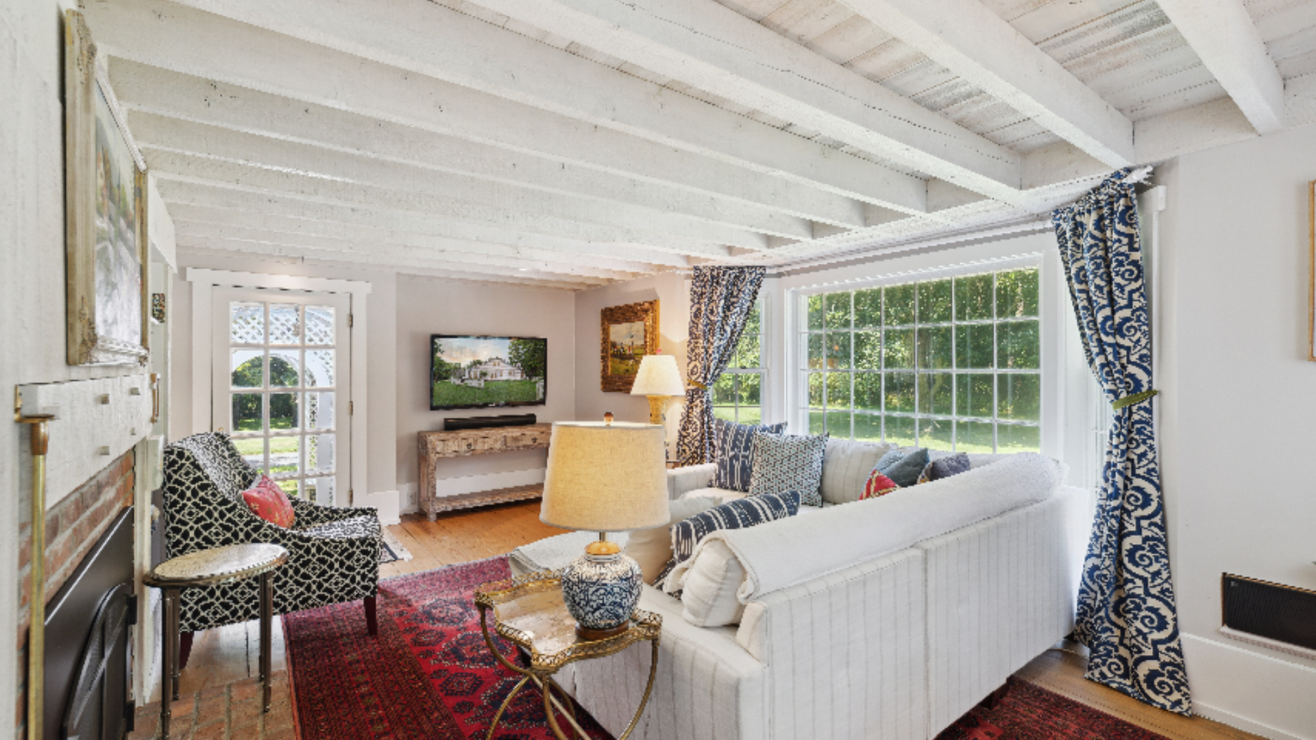 Beamed Cottage Living Room - White-painted exposed beams and a fireplace with a mounted TV create a cozy focal point, while a soft linen white sofa and patterned accent chairs offer relaxed, comfortable seating. Bold indigo drapery frames a wide wind