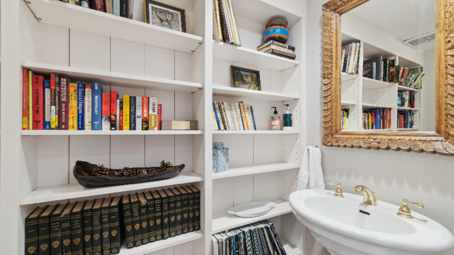 Bookcase Powder Room - A clever built-in bookshelf lends unexpected character to this powder room, filled with well loved books and collected treasures like a miniature model boat. A gold-framed mirror adds a touch of glamor above the pedestal sink, 