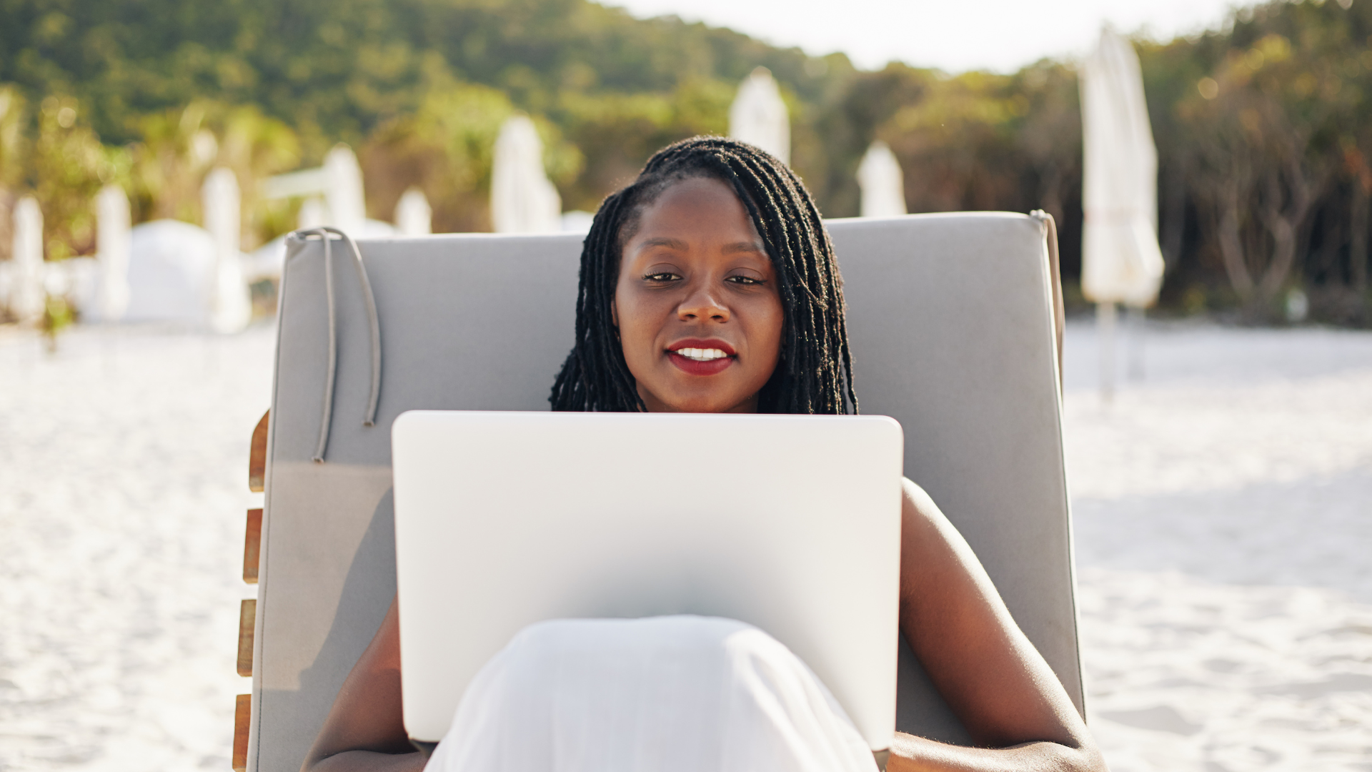 Femme assise sur une chaise de plage, regardant un ordinateur portable, plage en arrière-plan