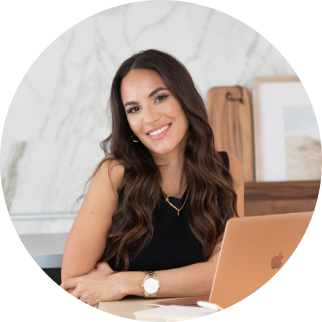 A young woman with long dark hair smiling at a desk with a laptop in a bright, modern office.