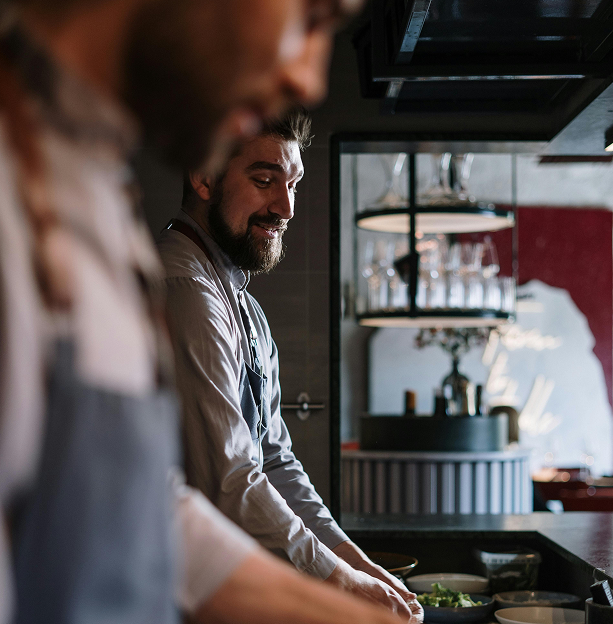 Two chefs preparing food in a restaurant kitchen, with shelves of glasses and a bar in the background.