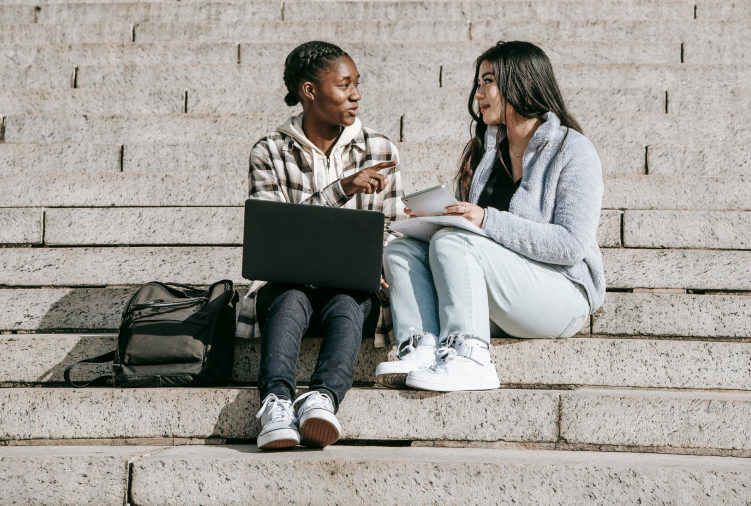 Two young women sitting on outdoor steps, having a conversation with a laptop, tablet, and backpack nearby.