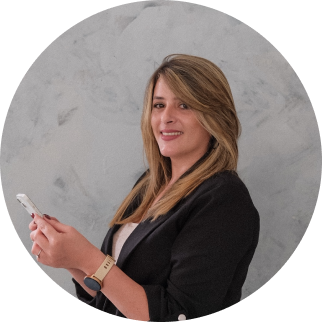 A woman with long blonde hair wearing a black blazer and a beige top, holding a smartphone and smiling at the camera, standing against a gray background.