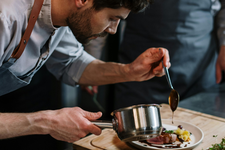 A chef tasting sauce with a spoon over a plated dish in a kitchen.