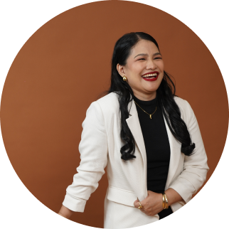 A woman with long black hair wearing a white blazer, black top, and gold jewelry, smiling against a brown background.