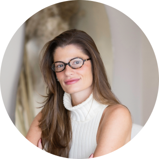 A woman with long brown hair, glasses, and a white sleeveless turtleneck sweater smiling at the camera.