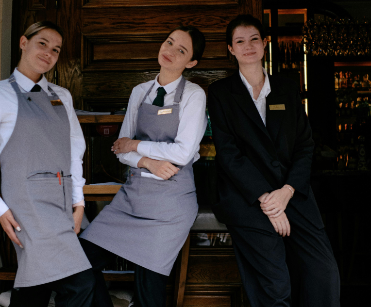 Three women working at a restaurant, wearing uniforms, inside a warmly lit dining area.