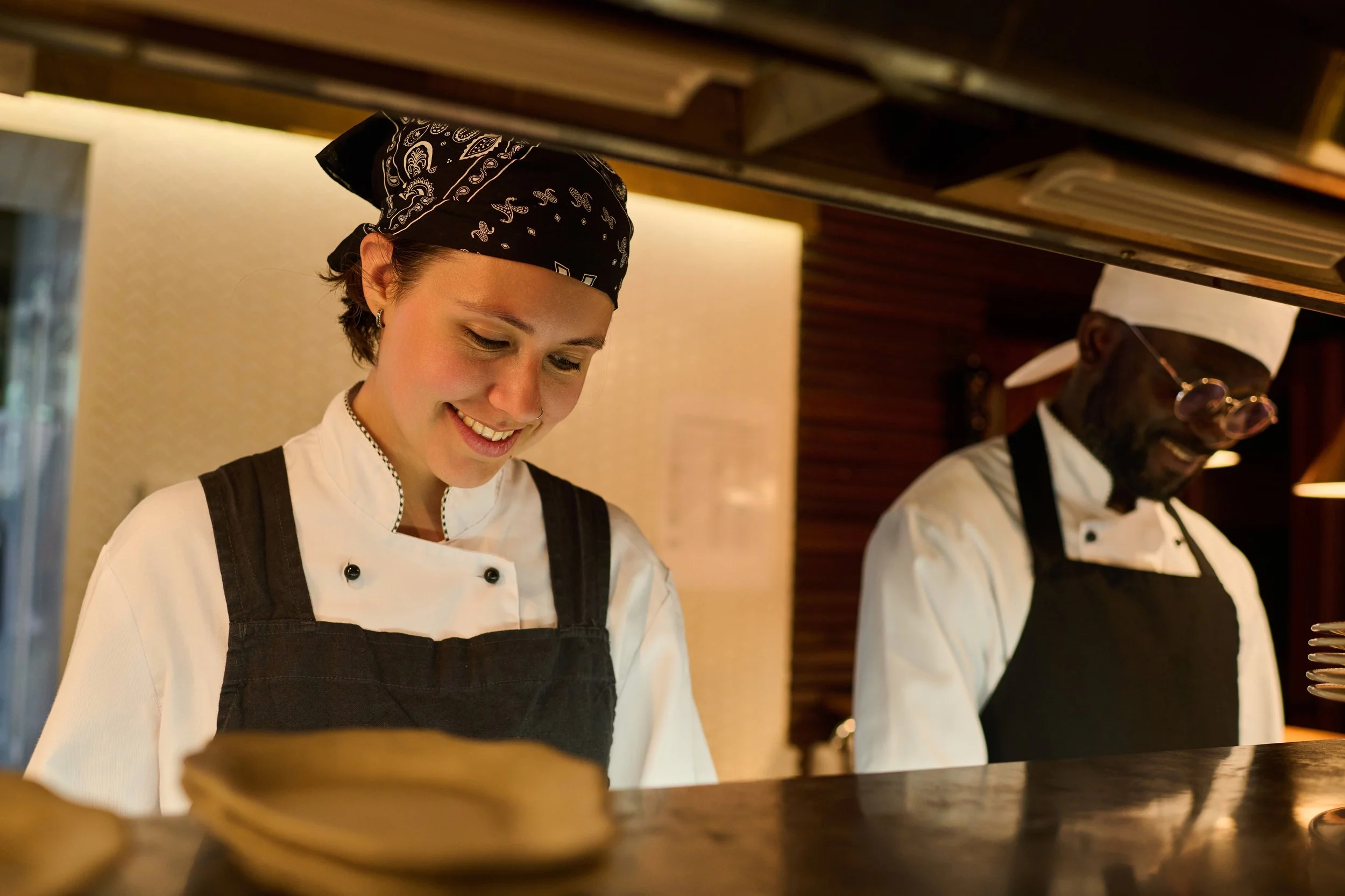 A female chef and a male chef working in a kitchen, both smiling and wearing chef uniforms and hats.