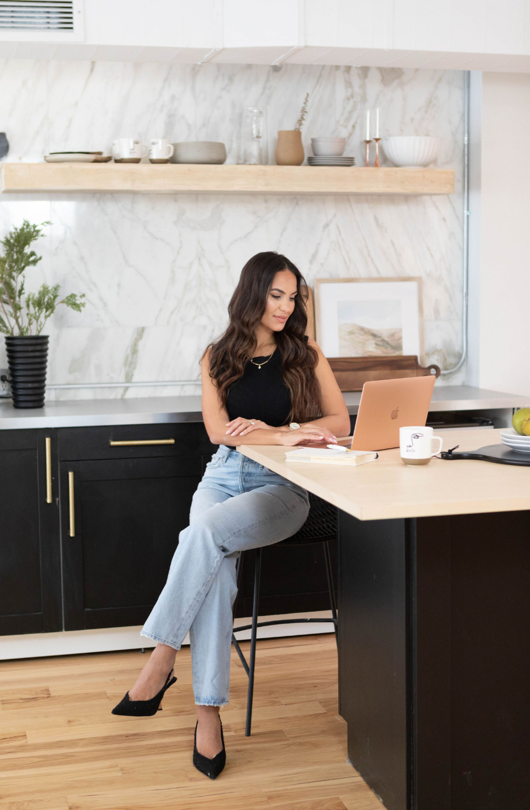 A woman working on a laptop at a kitchen island with a cup, a stack of books, and a plate of apples