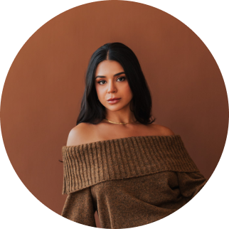 Portrait of a young woman with dark hair wearing an off-the-shoulder brown sweater, standing against a brown background.
