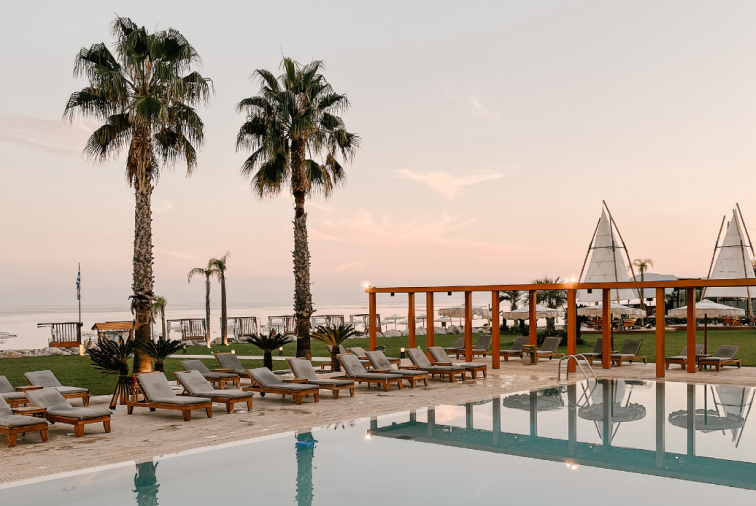 Swimming pool area with lounge chairs, palm trees, a wooden pergola, and beach umbrellas in the background during sunset.