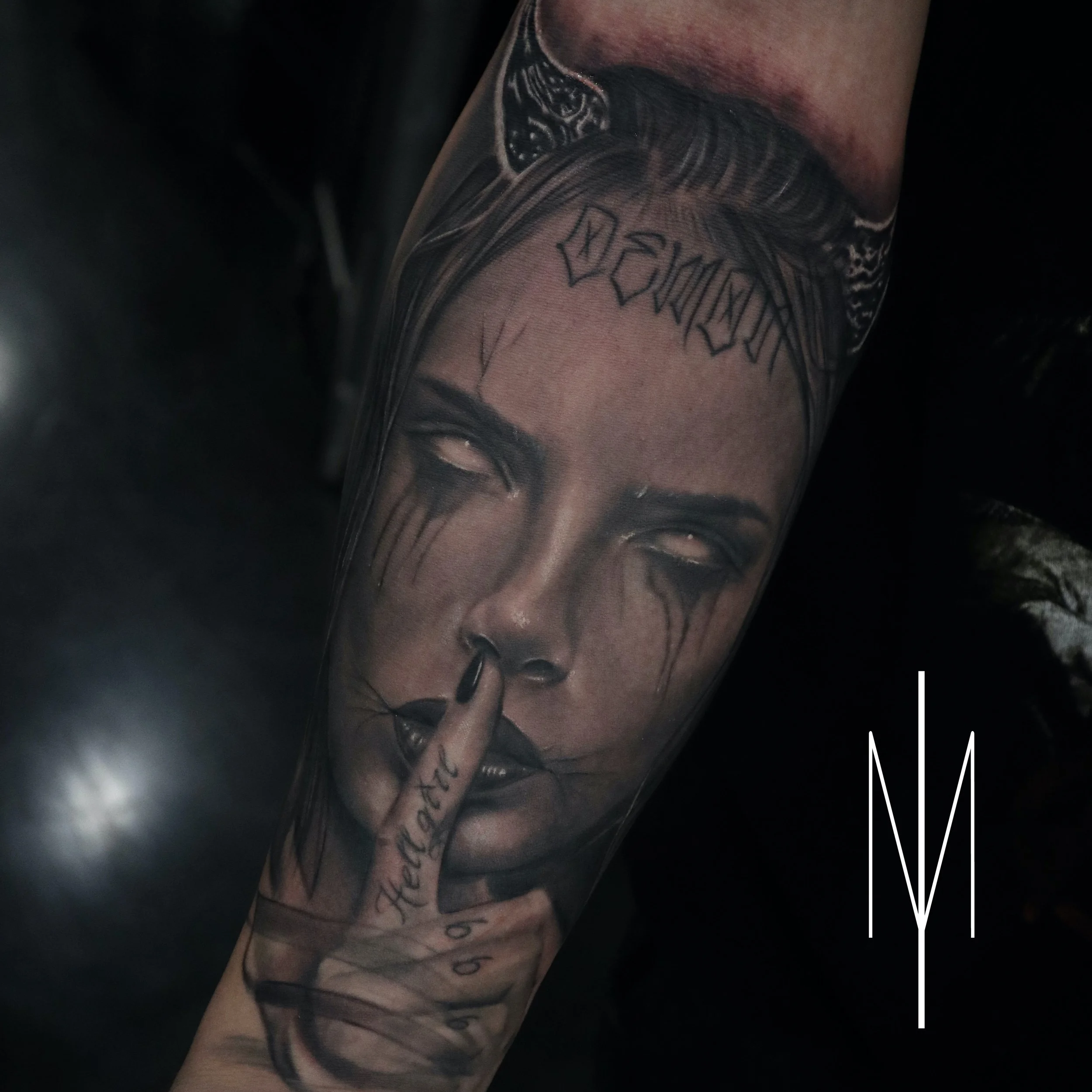 Tattoo of a woman's face with closed eyes and black tears streaming down her cheeks, with her finger placed on her lips in a shushing gesture. The Word "Demon" tattooed above her forehead and additional decorative elements are also visible.