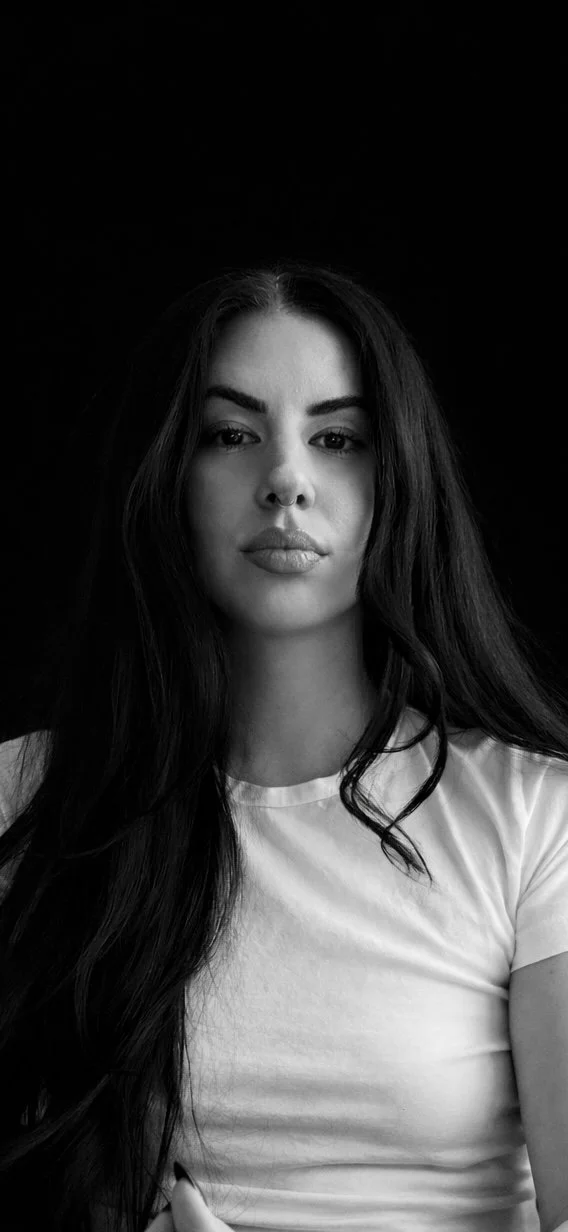 Black and white portrait of a young woman with long dark hair wearing a white T-shirt, against a dark background.