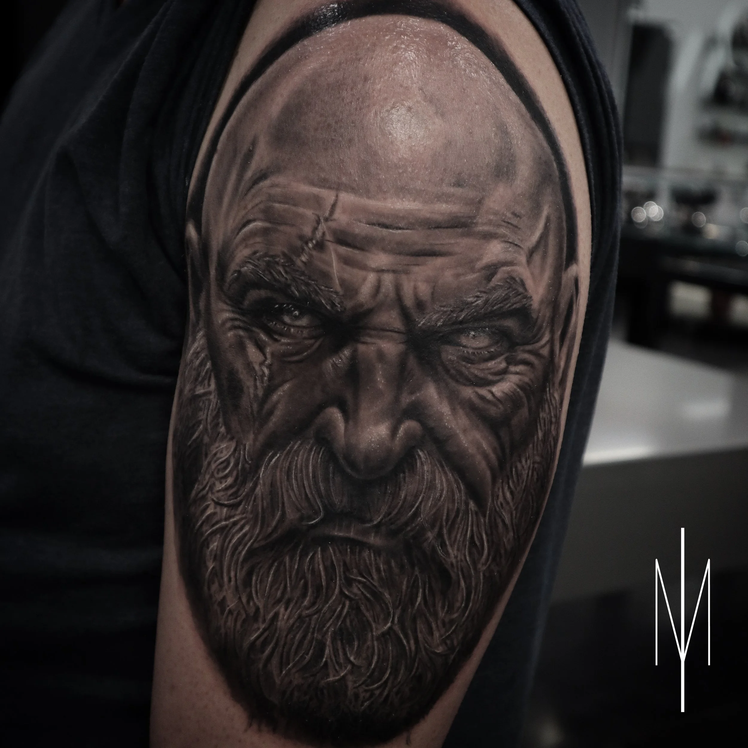 Detailed black and gray tattoo of the God of War with a beard, bald head, and intense expression on upper arm.