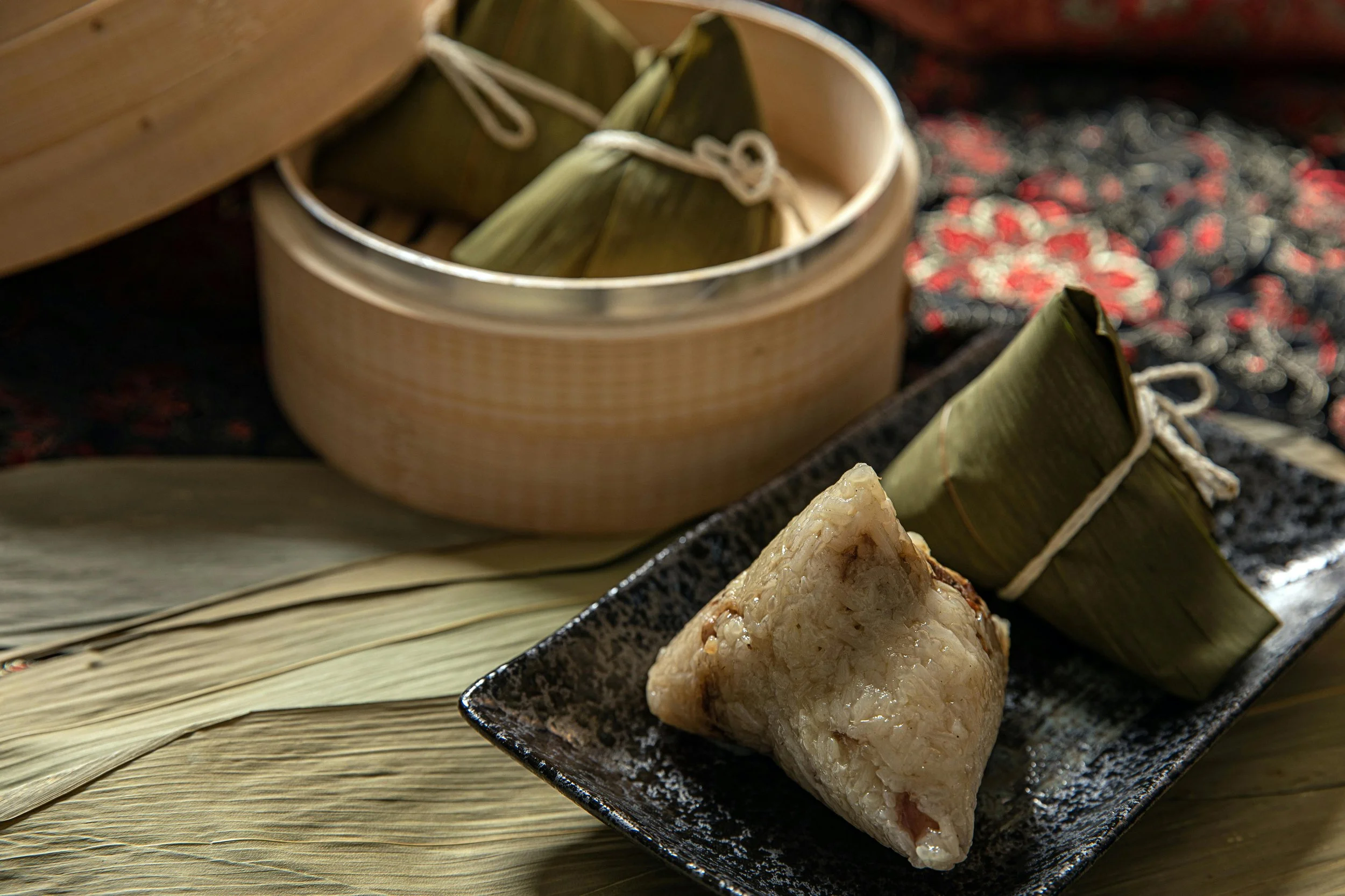 Tamale wrapped in corn husks and a Japanese rice dumpling on a black plate, with a bamboo steamer in the background.