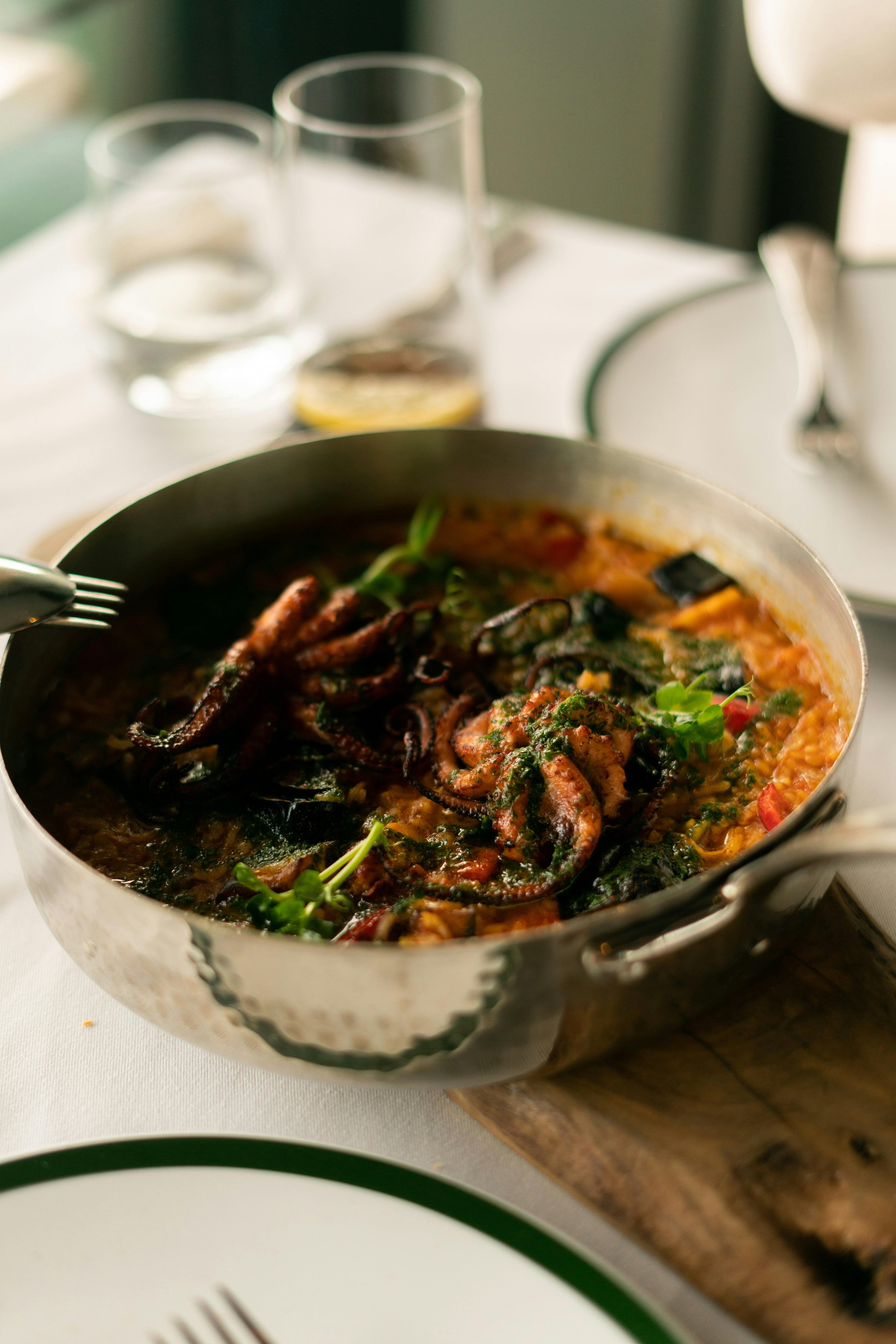 A seafood stew with octopus and greens served in a metal pot on a wooden trivet.