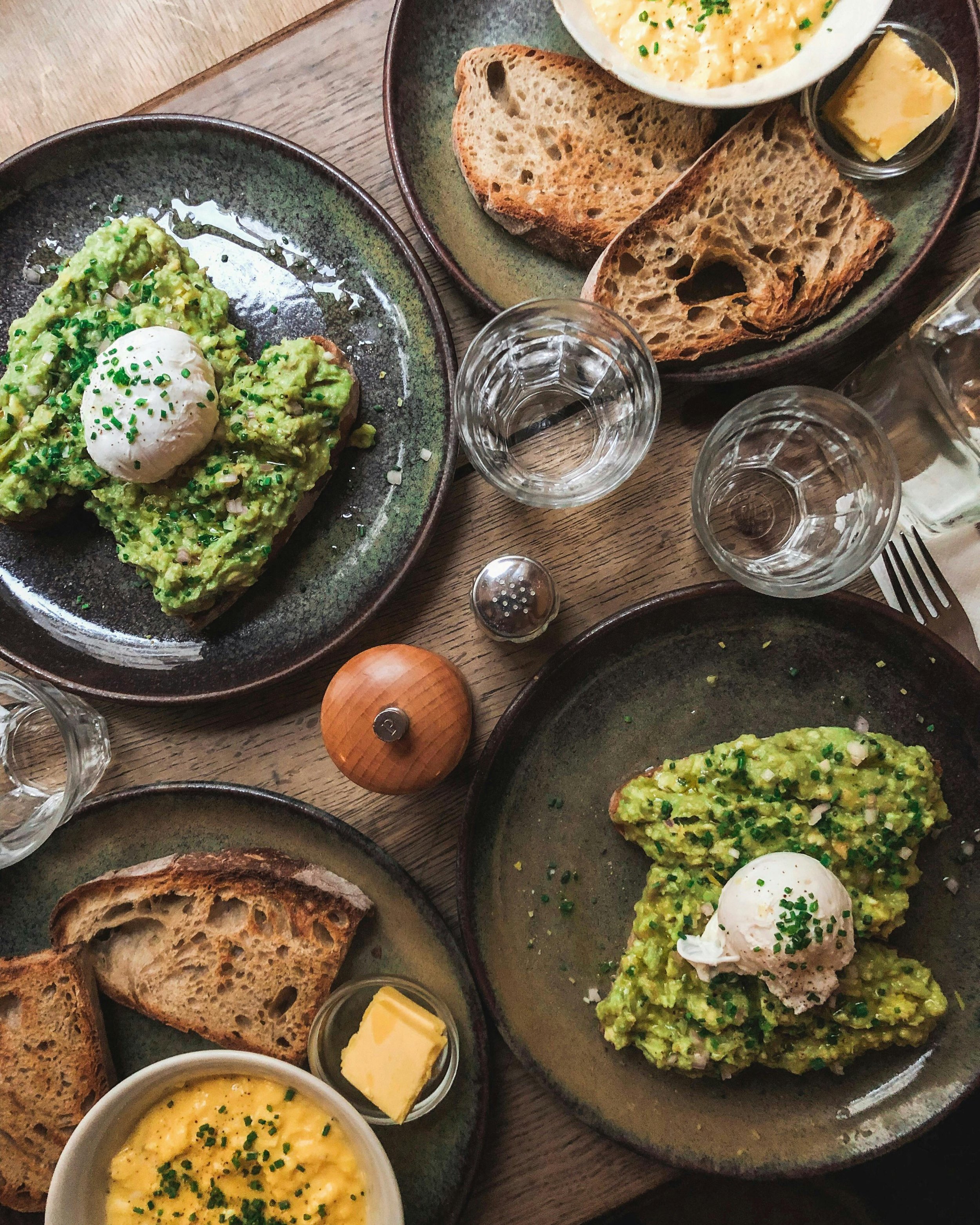 Plate with avocado toast topped with a poached egg and chives, slices of toasted bread, scrambled eggs with chives, butter, water glasses, and pepper shaker on a wooden table.