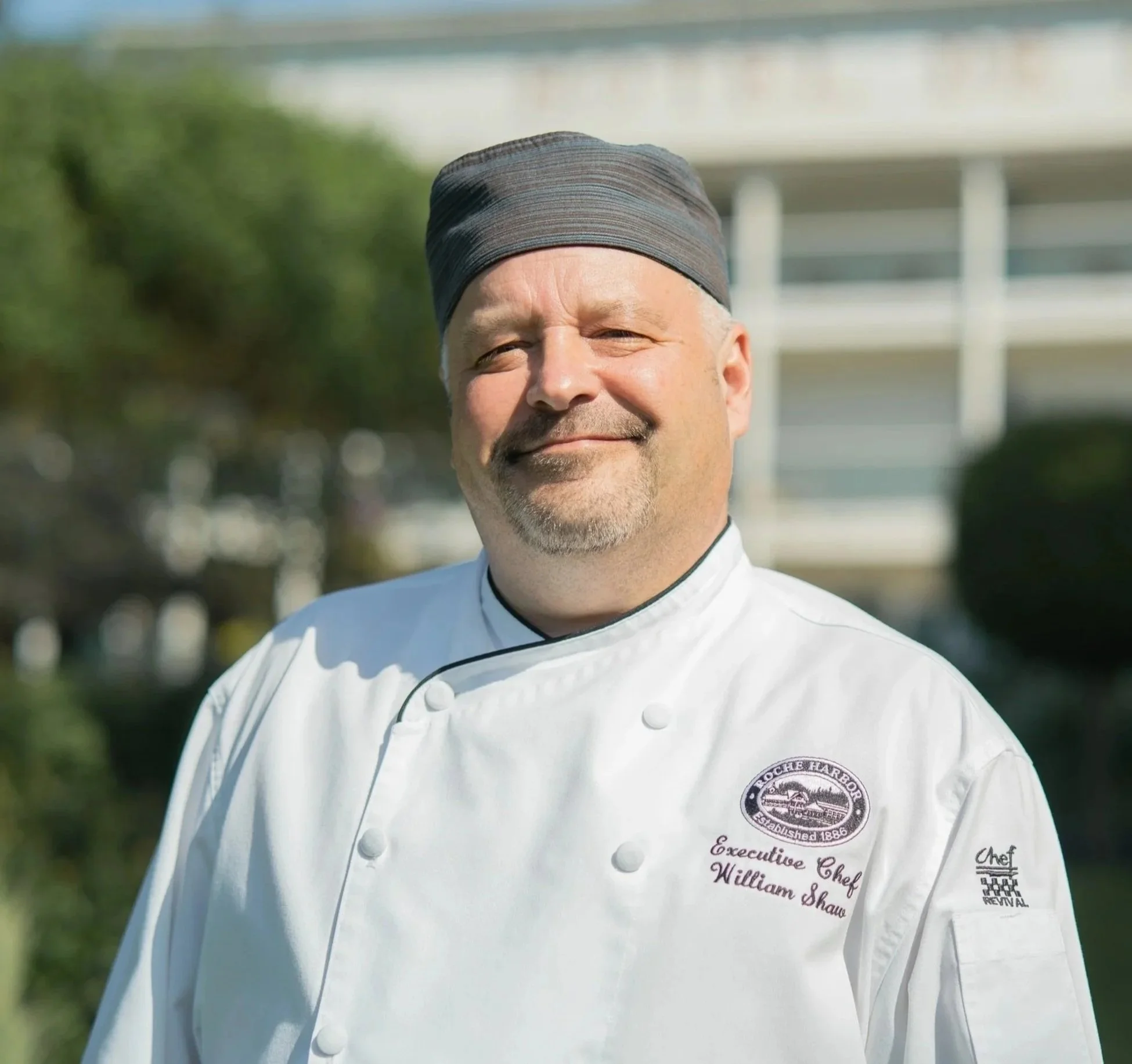A male chef wearing a white chef's coat and a black chef's hat, standing outdoors with greenery and a building in the background.