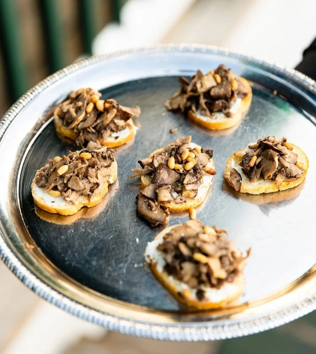 Small appetizers with toasted bread rounds topped with shredded meat and pine nuts on a silver tray.