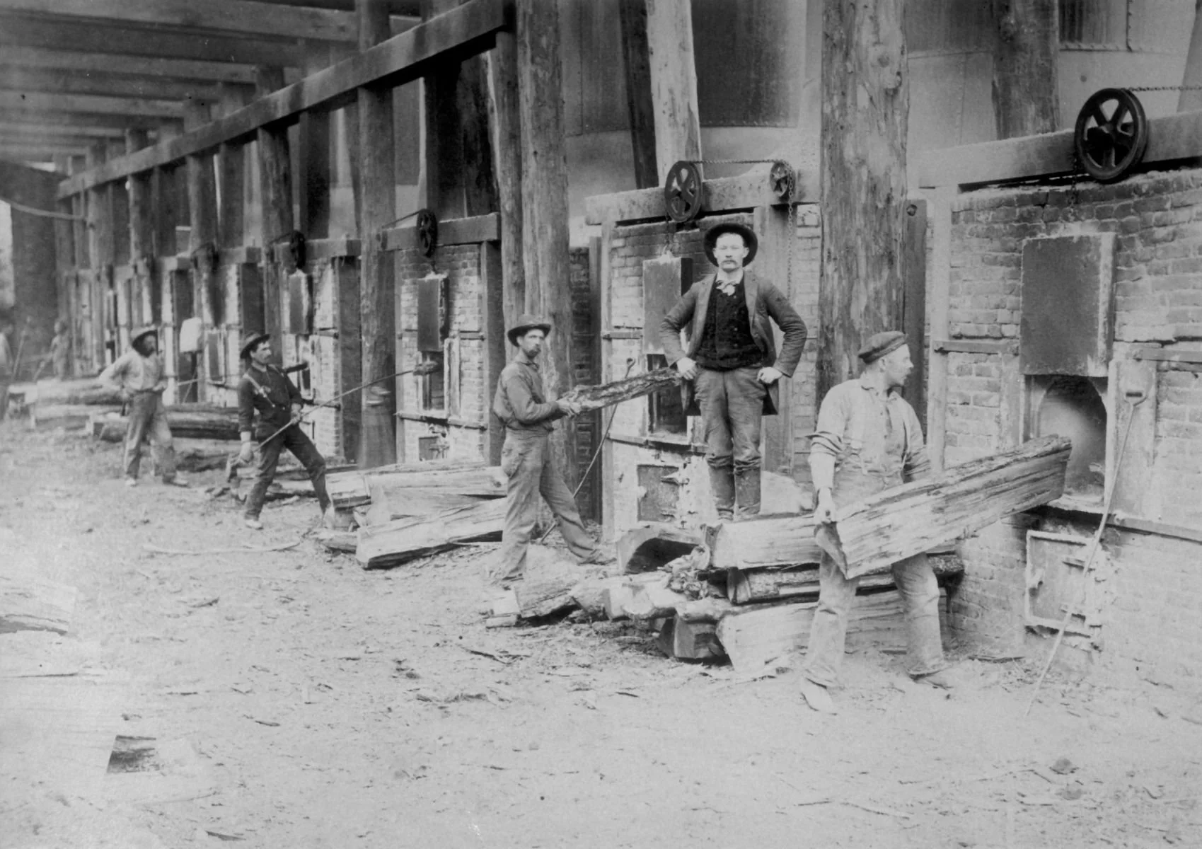 Image of several workers, some with hats, constructing a brick building with wooden beams and machinery, using large wooden planks and tools, during a historical time period.