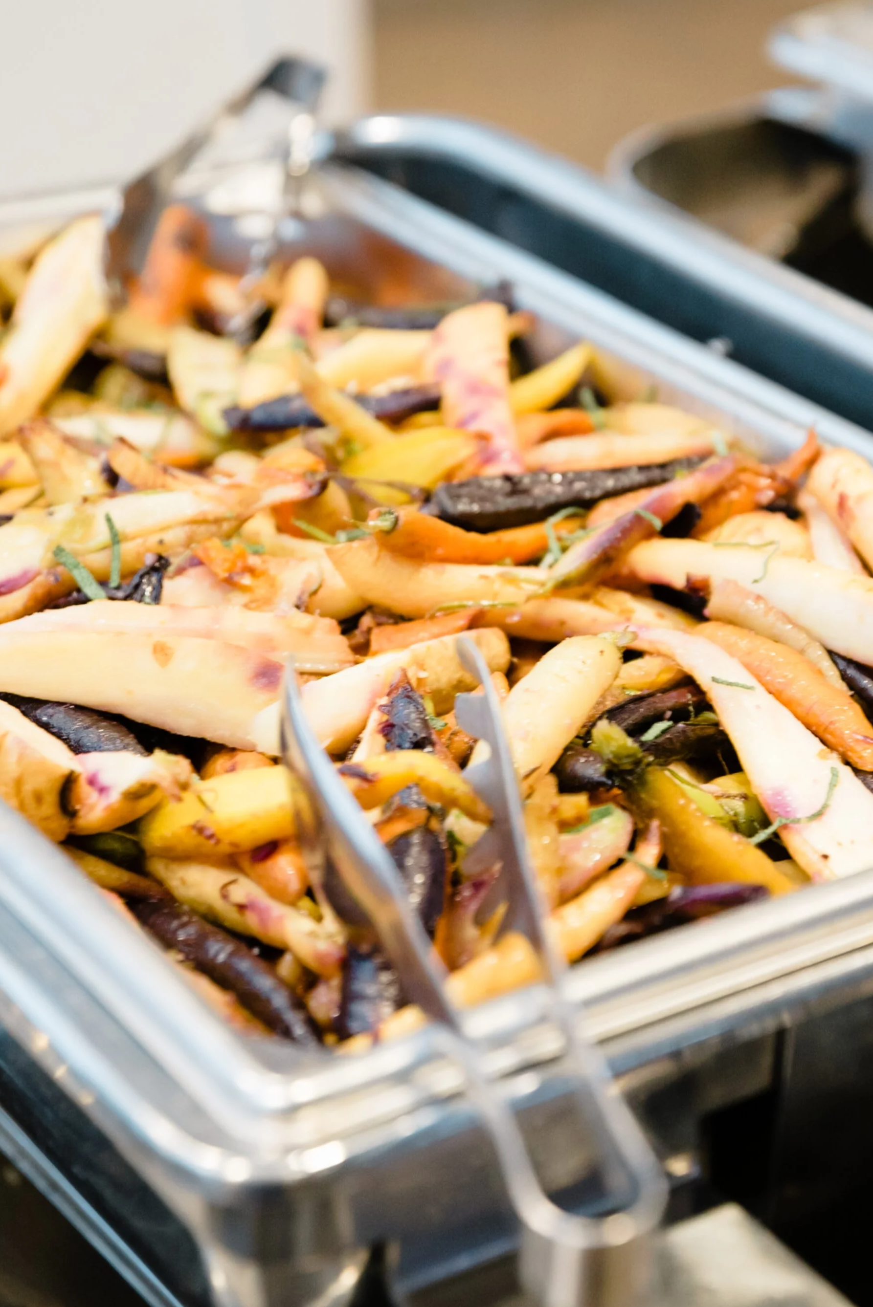 Close-up of a tray of roasted mixed vegetables including eggplant, carrots, and other root vegetables, with tongs resting on the tray.