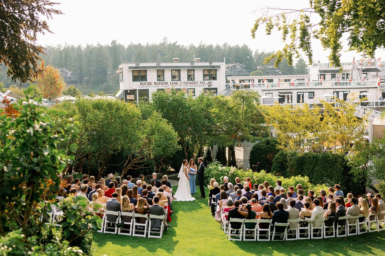 An outdoor wedding ceremony in a lush garden with a bride, groom, and officiant standing under trees, surrounded by seated guests on white chairs, with a white multi-story building in the background under a clear sky.