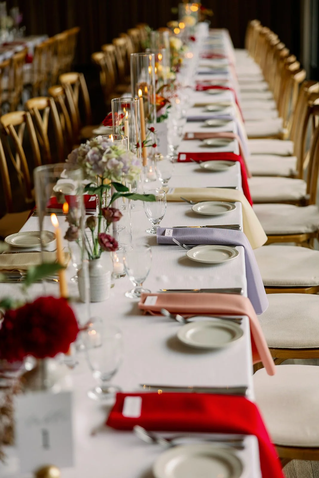 Long banquet table set for a formal event with floral centerpieces, candles, wine glasses, plates, and silverware, decorated with colorful napkins.
