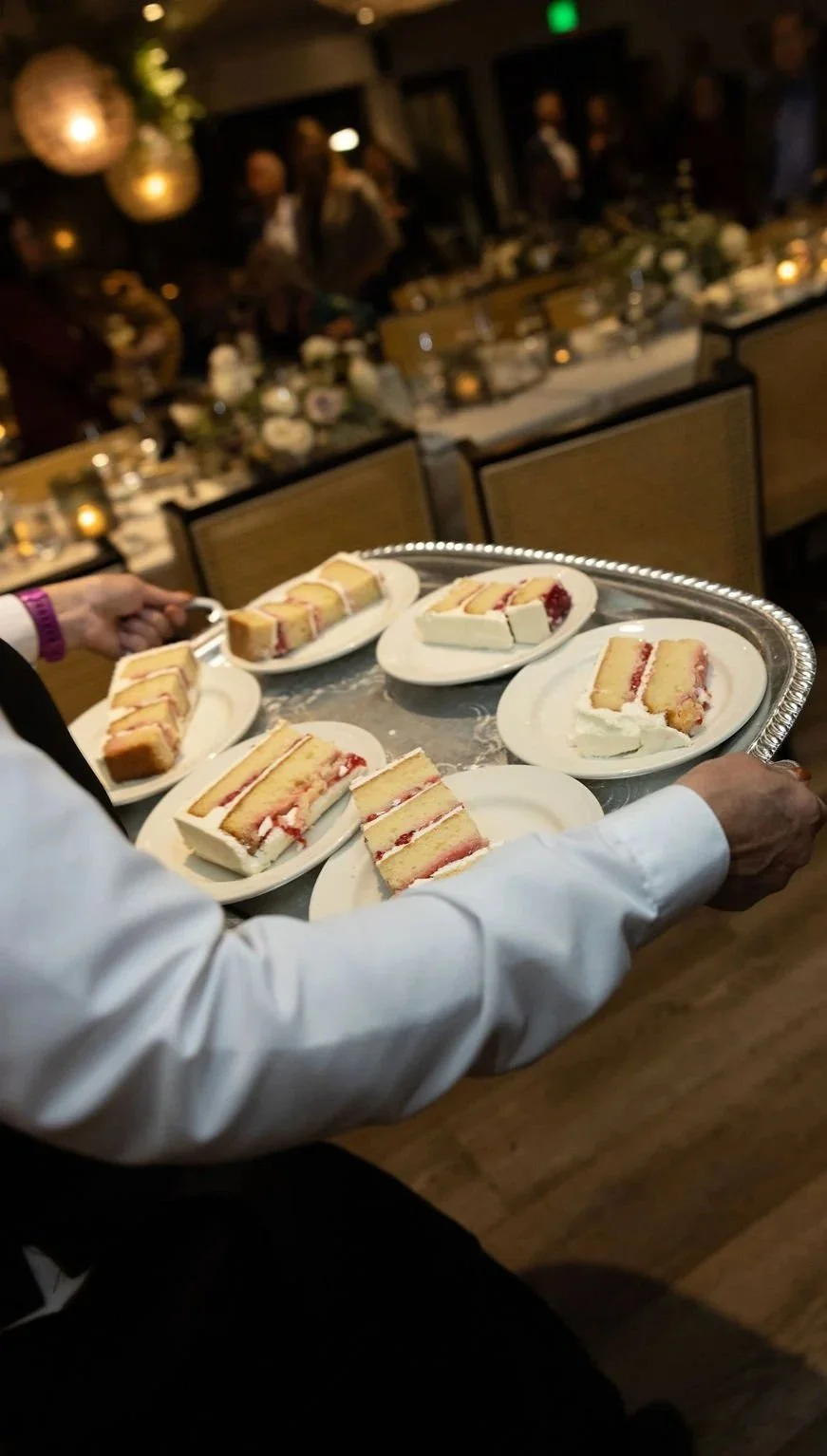 Person holding a silver tray with plates of sliced cake, set in a dimly lit dinner hall with decorated tables and hanging lights.