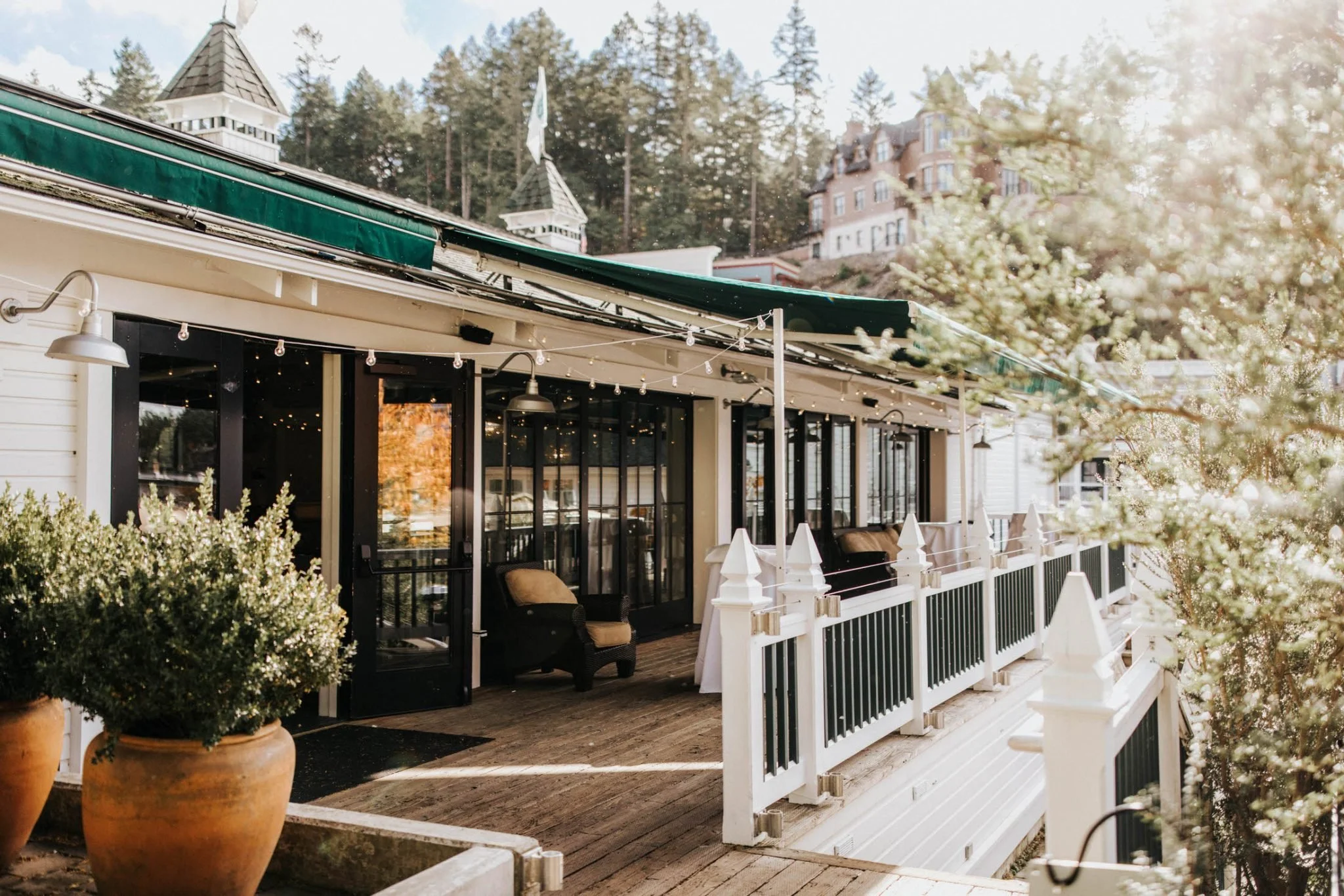 Exterior of a café or restaurant with large glass doors, outdoor seating, and string lights, with a hillside and buildings in the background.