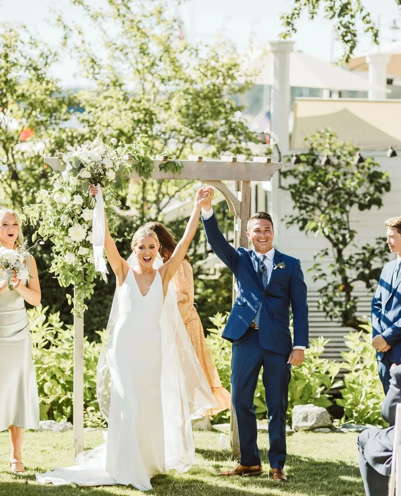 A newly married couple celebrating at their outdoor wedding, with the bride raising her bouquet and the groom holding her hand, under a decorated arch, surrounded by friends and greenery.