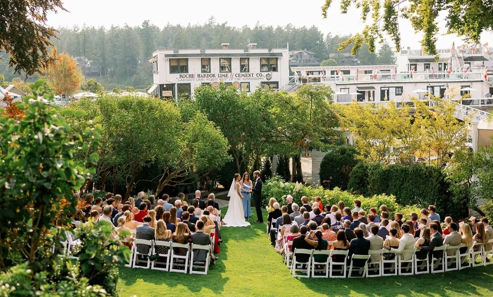 Outdoor wedding ceremony with a bride and groom exchanging vows under trees, surrounded by seated guests, with a white building and river in the background.