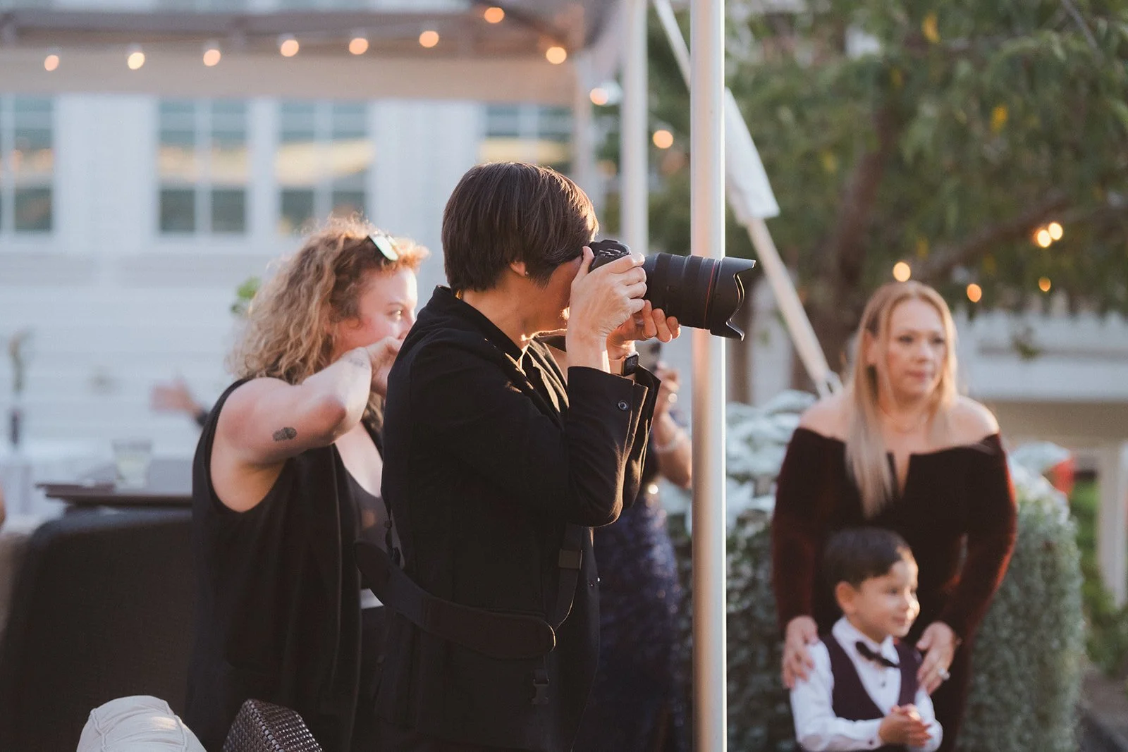 A photographer taking photos at an outdoor event during sunset, with guests including a woman with curly hair and a woman with straight blonde hair holding a young boy in a tuxedo.
