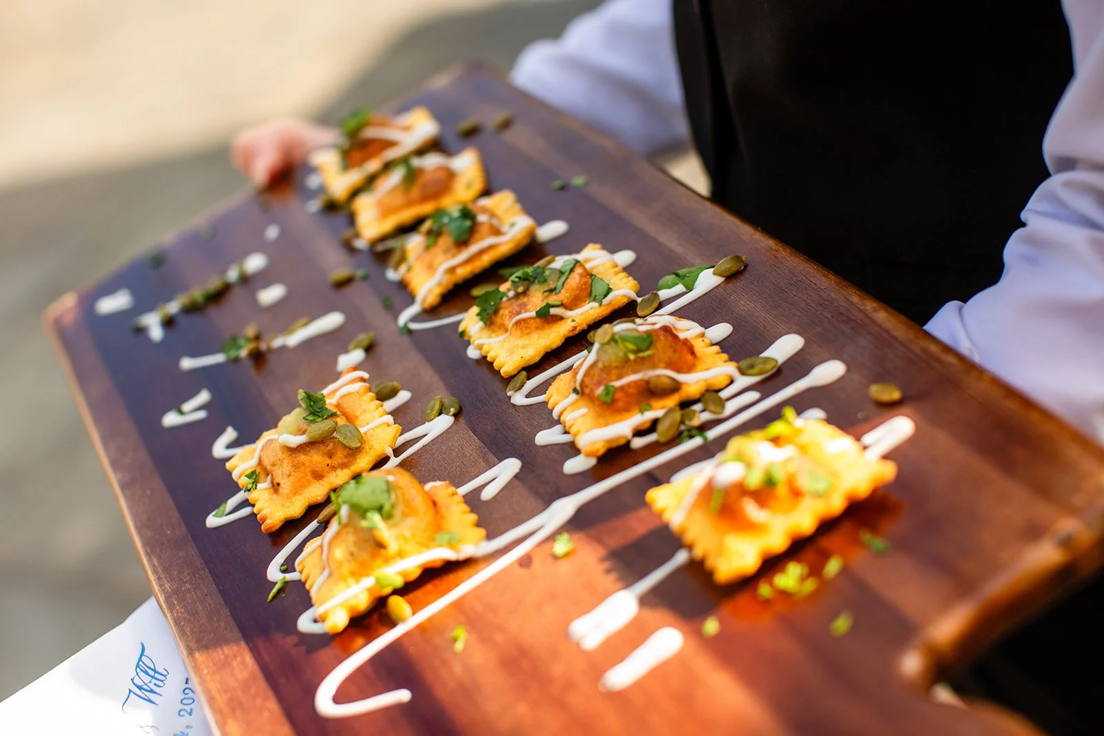 A wooden serving board with several pieces of ravioli pasta topped with chopped herbs, pumpkin seeds, and drizzled with white sauce.