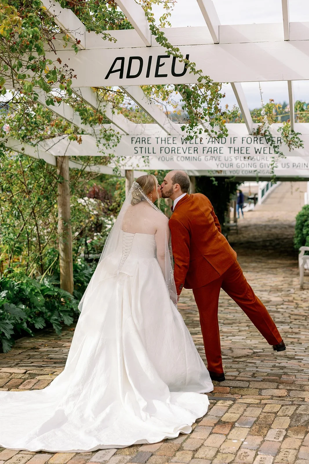 A bride and groom sharing a kiss under an archway with a sign that says 'ADIEU'. The bride wears a white wedding dress with a long train and a veil, and the groom wears a rust-colored suit. They are standing on a cobblestone path surrounded by greenery.