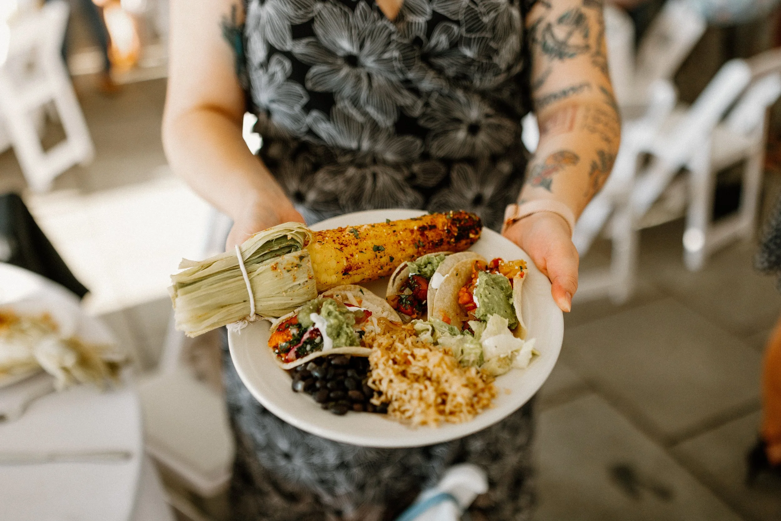 Person holding a plate of Mexican food with corn on the cob, tacos, black beans, rice, and a bundle of corn husks.