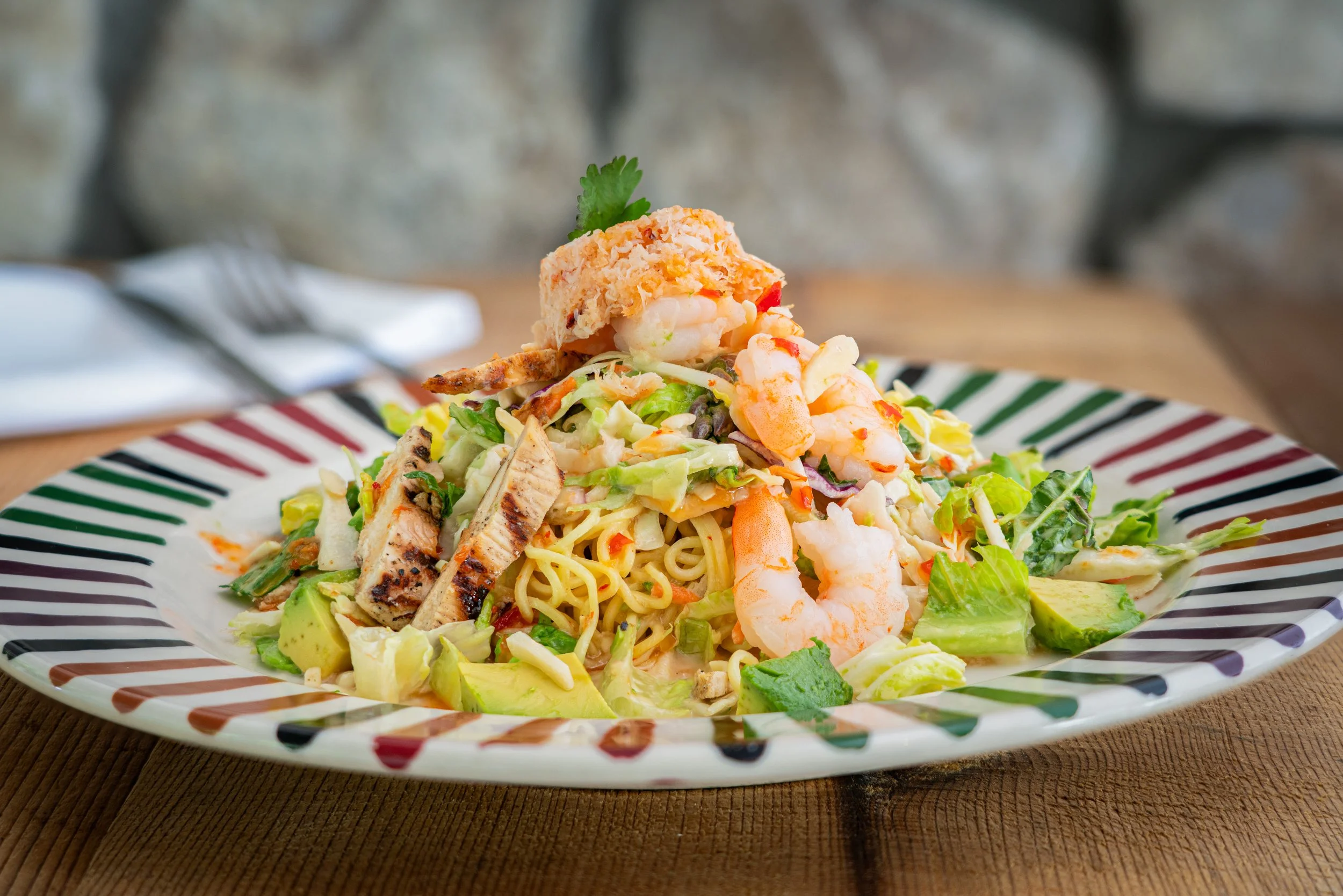 A plate of seafood pasta salad with shrimp, grilled chicken, vegetables, and a crab cake, garnished with cilantro