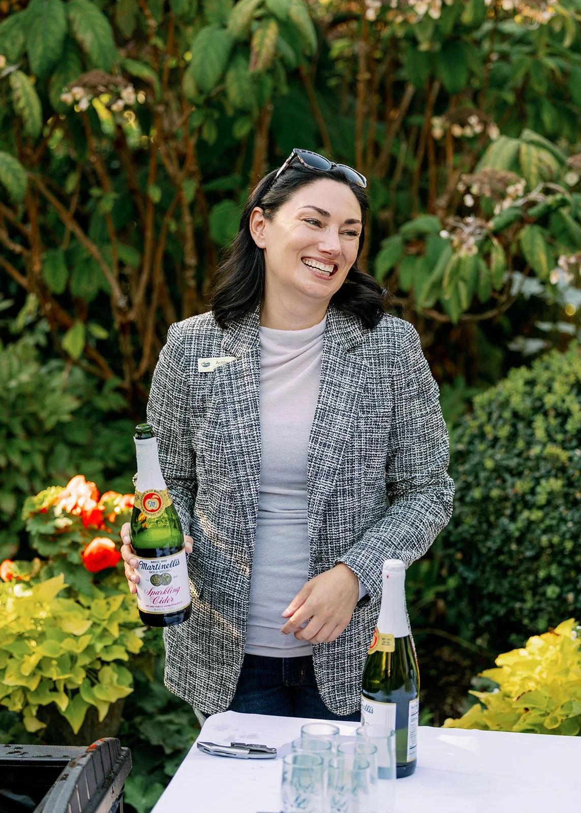 A woman smiling outdoors holding a bottle of sparkling cider at a table with glasses, with greenery in the background.