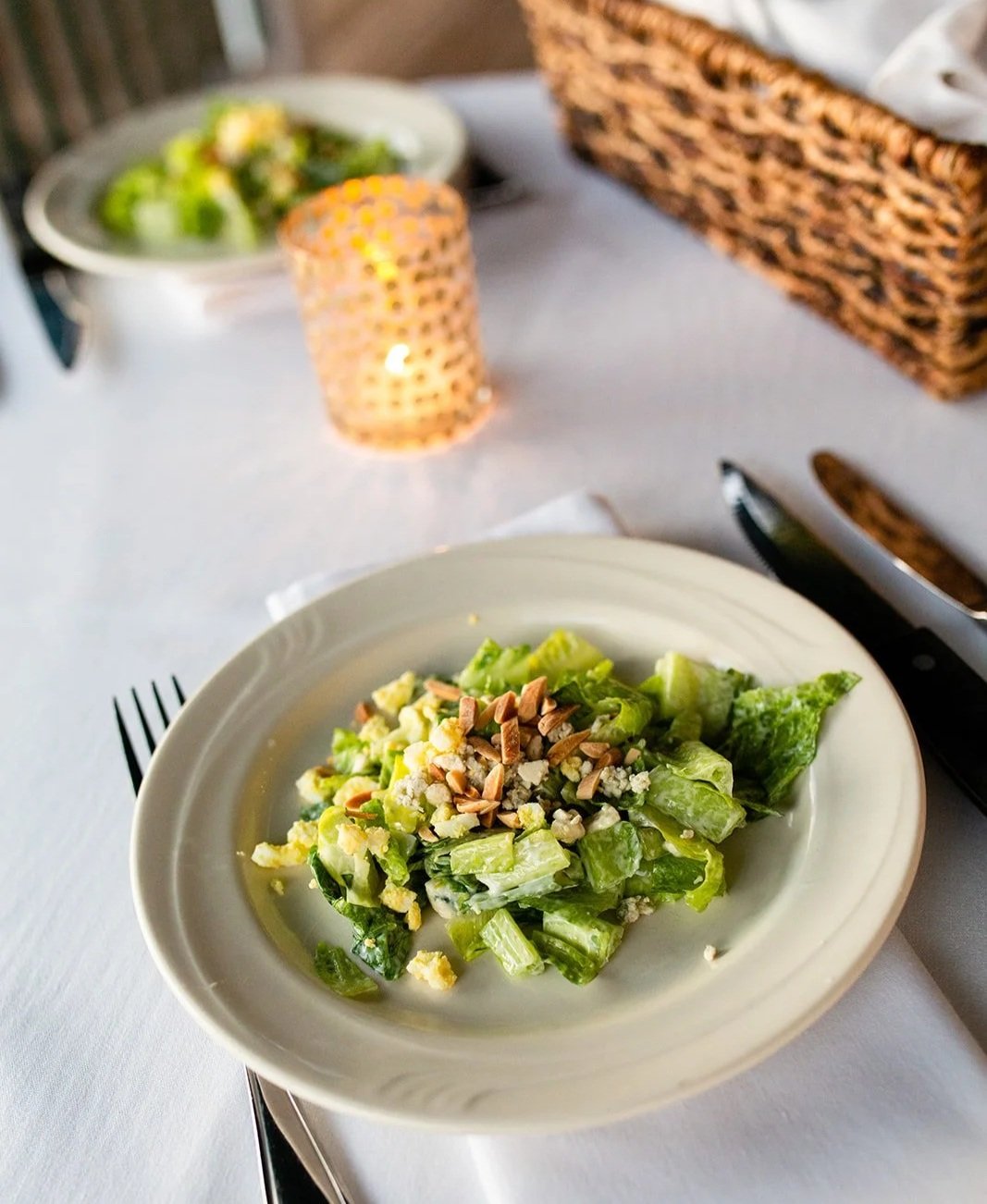 A plate of Caesar salad topped with shredded cheese and chopped nuts, on a white tablecloth with a fork, knife, and a candle in the background.