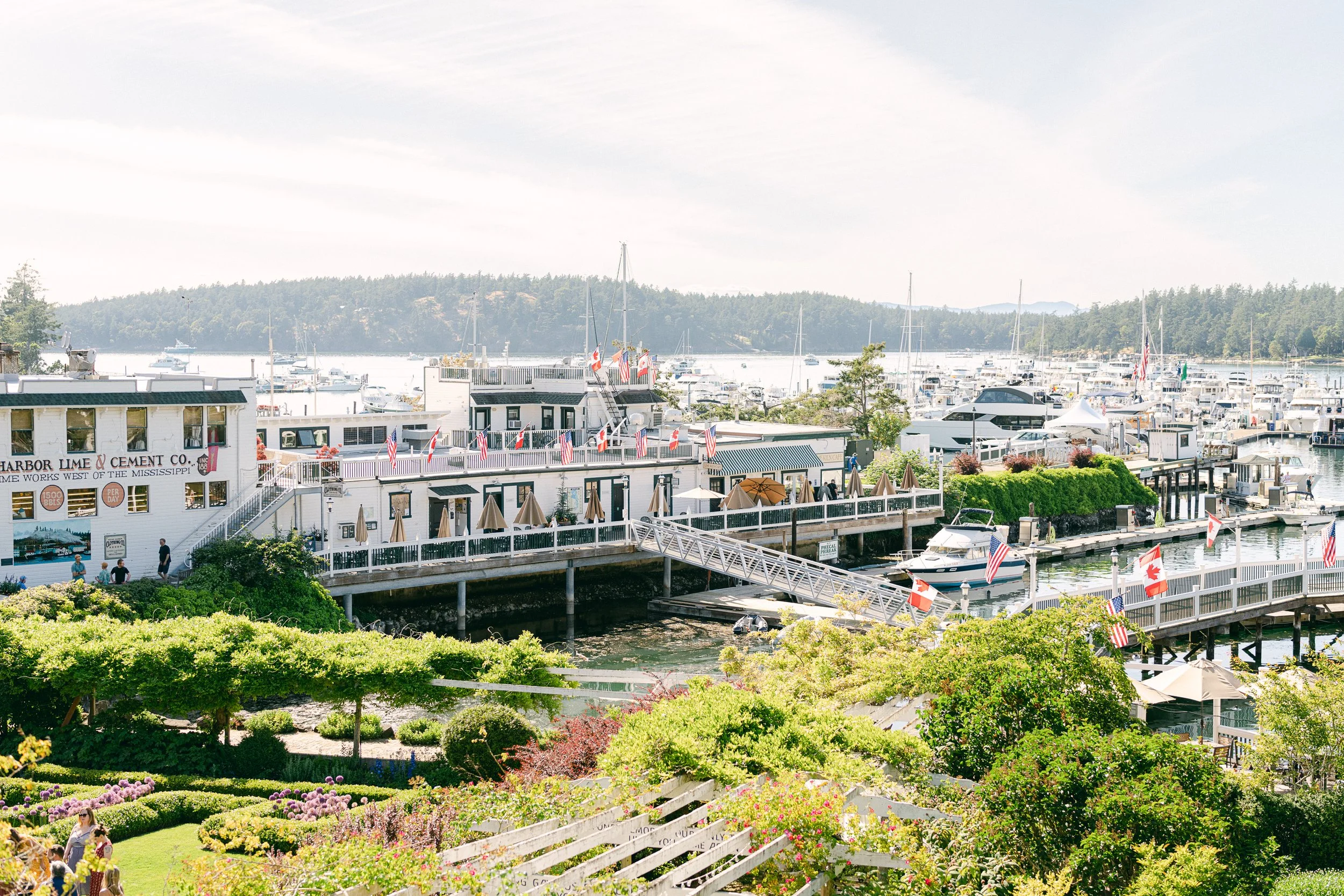 A scenic harbor with boats docked, a white building with a sign for Lime & Cement Co., and a garden with colorful flowers and trees in the foreground.