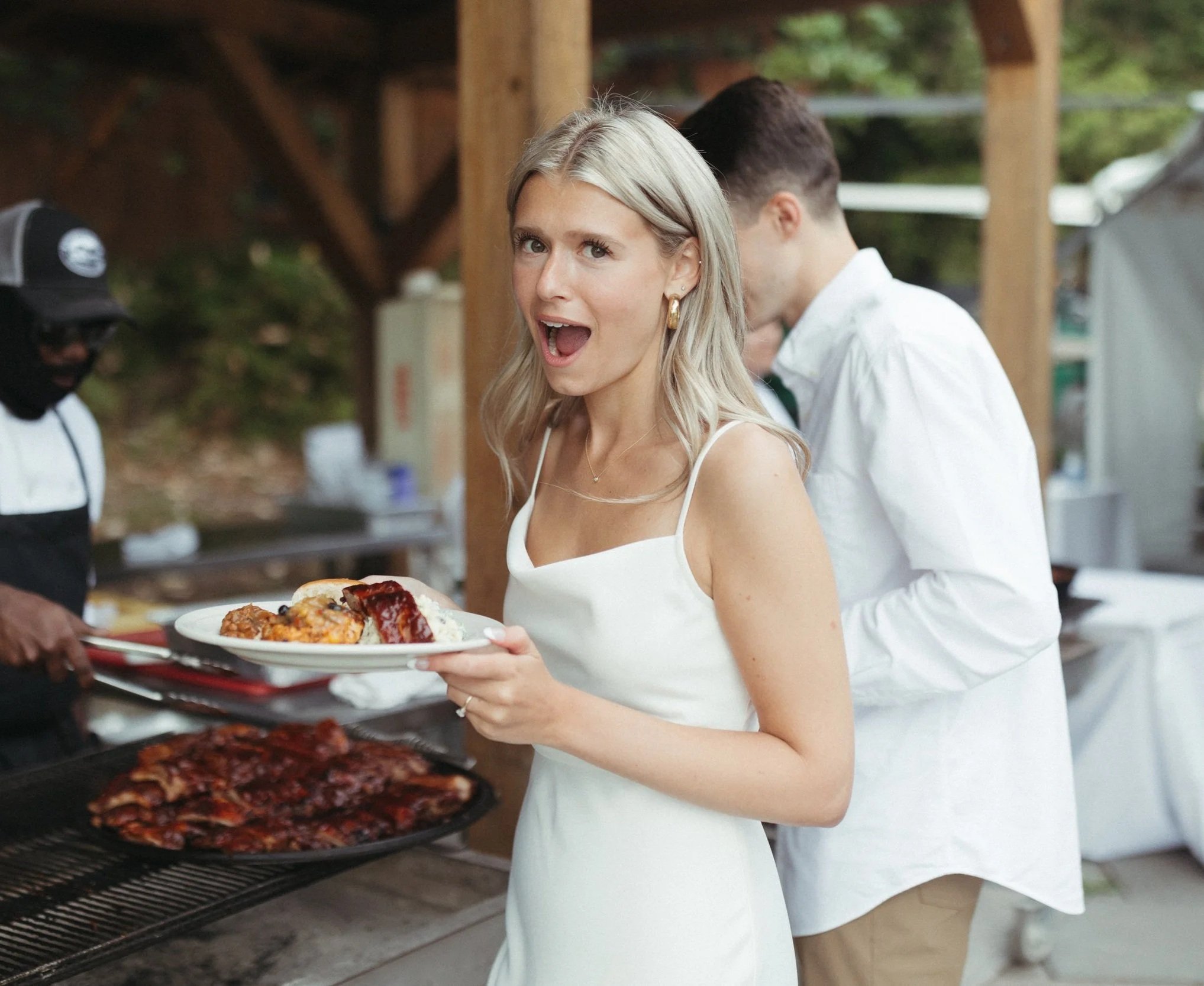 A woman holding a plate of food at an outdoor barbecue, smiling with surprise while standing near a grill with ribs, in a casual outdoor setting.