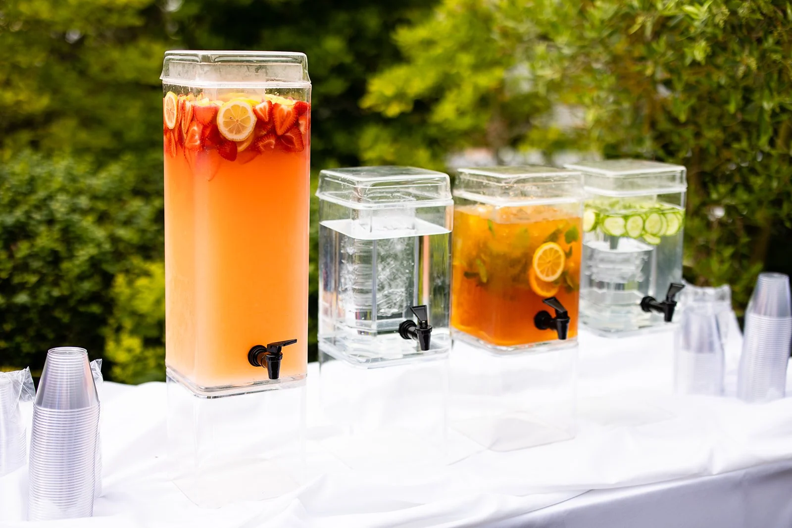 Arrangement of four large glass beverage dispensers with spigots on a table, filled with colorful fruit-infused water: orange-pink, clear, orange-yellow, and clear with cucumber slices, set against a background of green foliage.